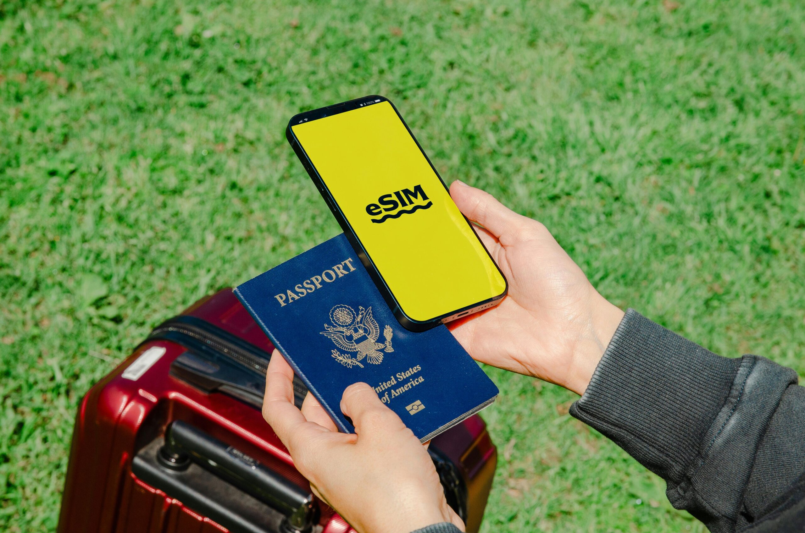 Hands holding a smartphone with eSIM and a passport near a luggage outdoors, ideal for travel concepts.